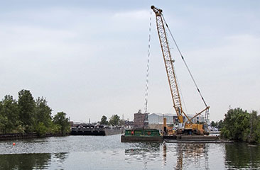 The dredging operation features a 230-ton crane that operates an environmental clam bucket. The bucket is submerged to the bottom of the waterway and then scoops up sediment. The dredged material is then loaded onto a hopper barge for transfer to the Confined Disposal Facility (CDF).