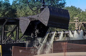 An excavator unloads dredged material into another hopper barge using a screen to separate the large debris. The sediment is mixed with recirculated water from the CDF to create a slurry.